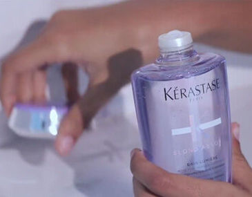 A close-up of a person's hand holding a bottle of Kerastase shampoo, with another product visible in the background.