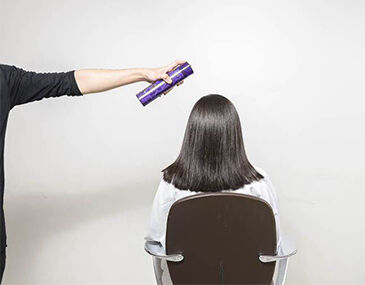 A person in a black shirt holds a hair spray can above the head of a seated individual with straight, dark hair, who is facing away from the camera. The background is a plain white wall.