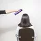 A person in a black shirt holds a hair spray can above the head of a seated individual with straight, dark hair, who is facing away from the camera. The background is a plain white wall.