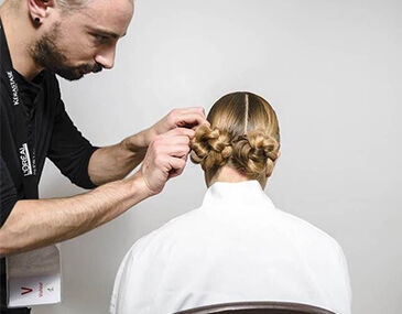 A hairstylist is styling a woman's hair into two elegant bun hairstyles from behind, with the stylist focusing on the details as she sits in a white chair.