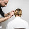 A hairstylist is styling a woman's hair into two elegant bun hairstyles from behind, with the stylist focusing on the details as she sits in a white chair.