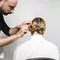 A hairstylist is styling a woman's hair into two elegant bun hairstyles from behind, with the stylist focusing on the details as she sits in a white chair.