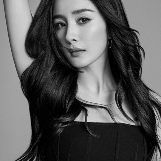A black and white portrait of a woman with long, flowing dark hair, wearing a strapless top and posing elegantly with one arm raised.