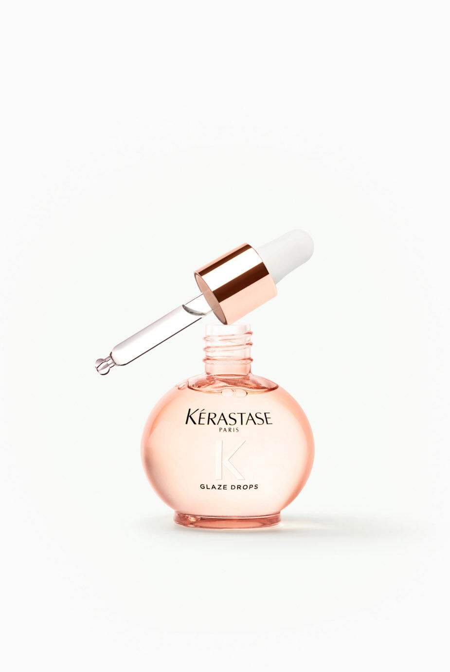 A round pink glass bottle of Kerastase Glaze Drops with a dropper cap, sitting upright with the dropper raised above the bottle.