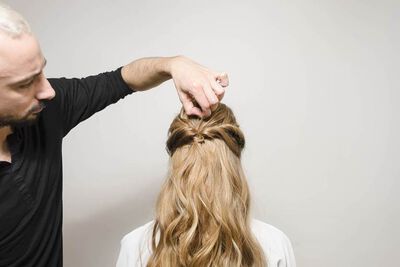 A hairstylist is creating a hairstyle on a person's long, wavy hair from behind, focusing on a section near the crown of the head.