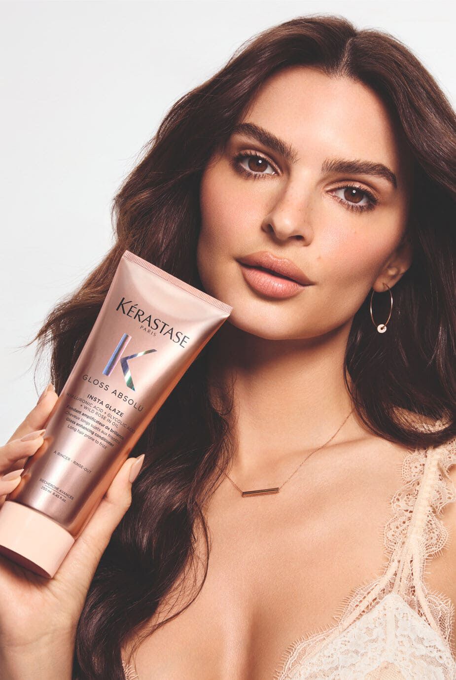 A woman with long, wavy hair holds a tube of Kerastase Gloss Absolu hair treatment, looking directly at the camera with a soft smile. She is wearing subtle jewelry and has a natural makeup look.
