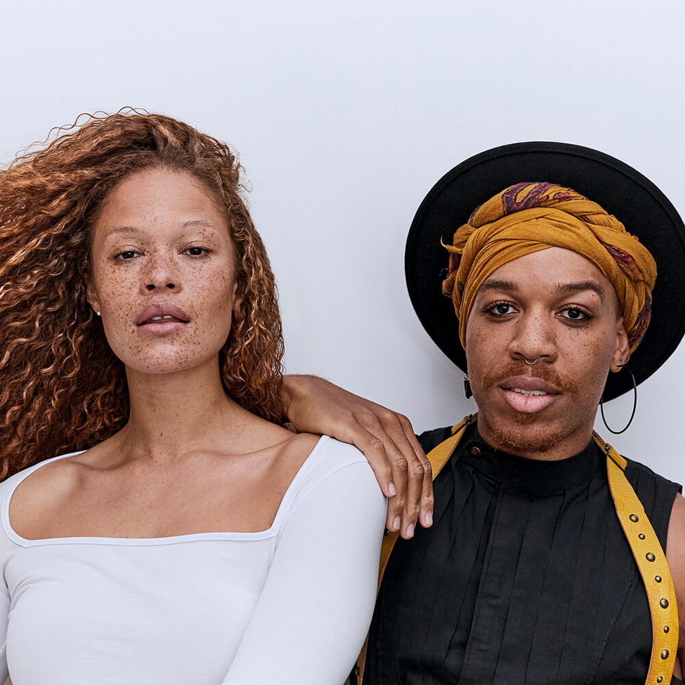 A woman with curly hair and a skin tone that shows freckles stands beside a man wearing a hat and a colorful headwrap. They pose together against a light background, conveying a sense of confidence.