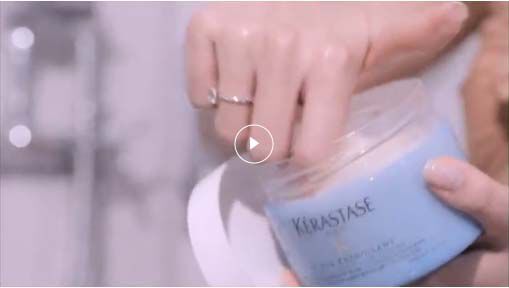 A hand reaching into a blue Kerastase hair mask container, highlighting the product in a spa-like setting.