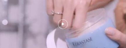 A hand reaching into a blue Kerastase hair mask container, highlighting the product in a spa-like setting.