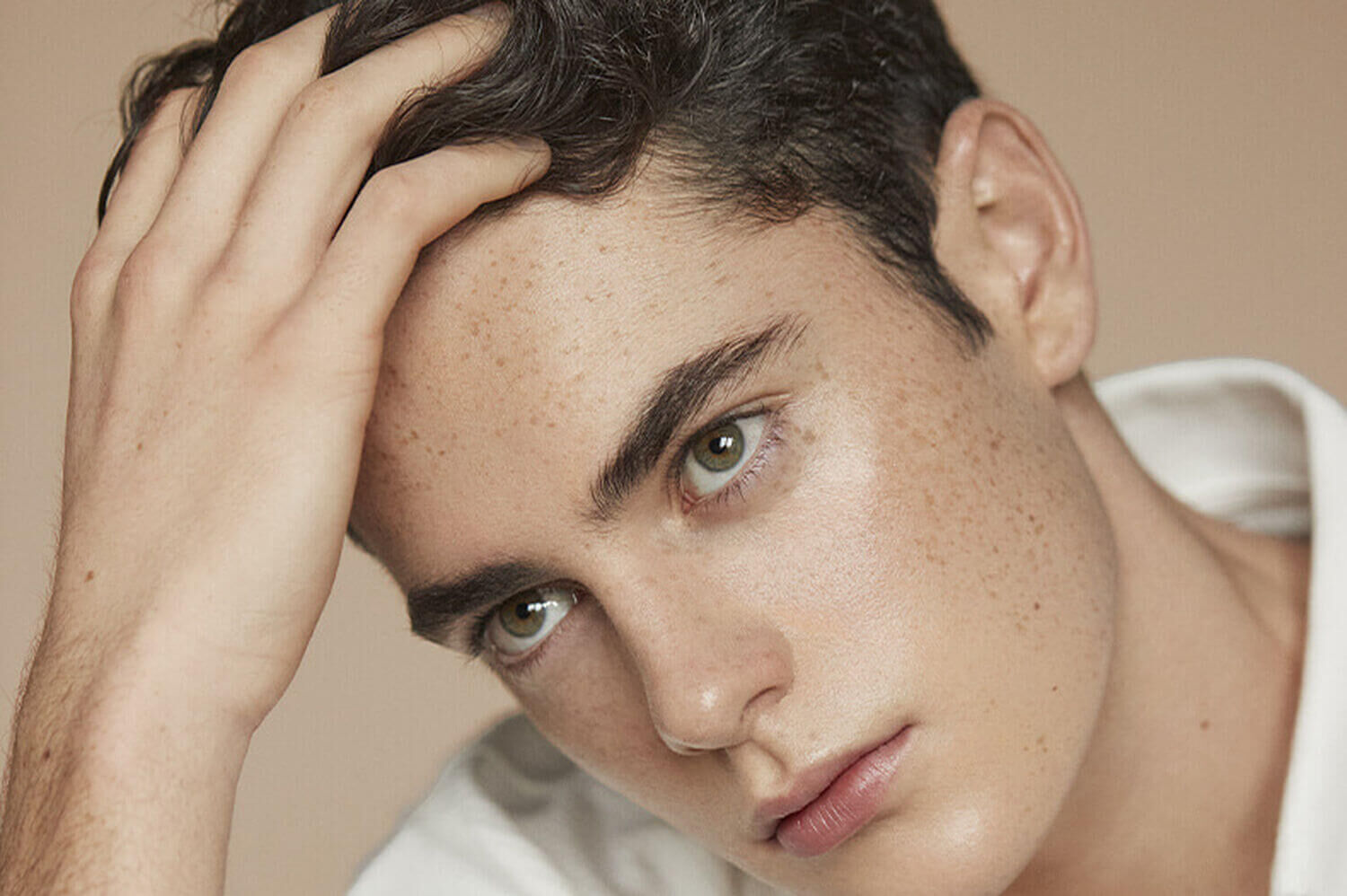 A young man with short dark hair and freckles poses thoughtfully, resting his hand on his head, against a neutral background.