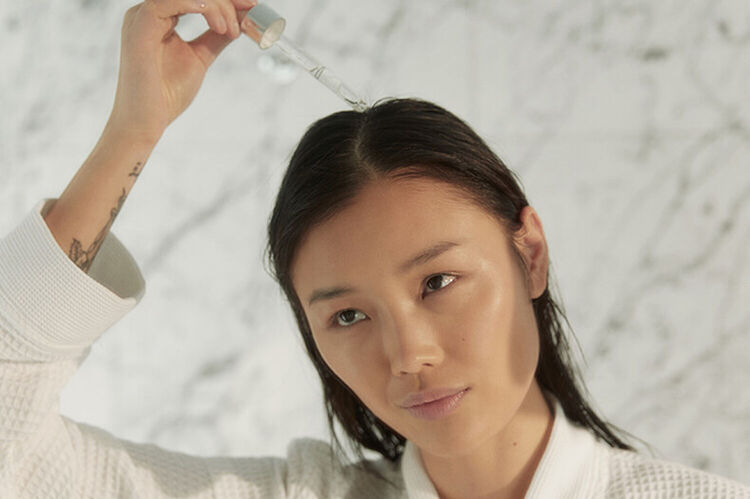 A woman in a white robe applies a dropper of serum to her hair, gazing at her reflection in a marble bathroom.