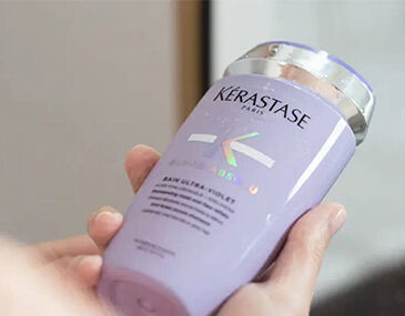 A person holding a bottle of Kerastase purple shampoo with a sleek design and holographic label.