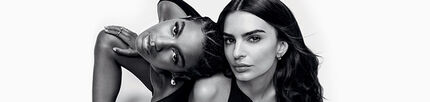 Two women pose closely together in a black and white photograph, both looking directly at the camera. One woman has short, textured hair and is positioned slightly behind the other, who has long, flowing hair. They both wear dark clothing and convey a sense of intimacy and connection.