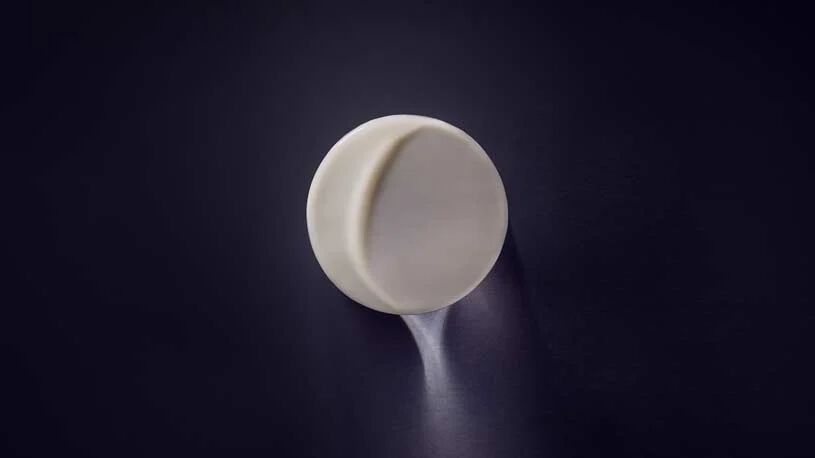 A minimalist, round, white pill rests on a dark surface, casting a soft shadow.