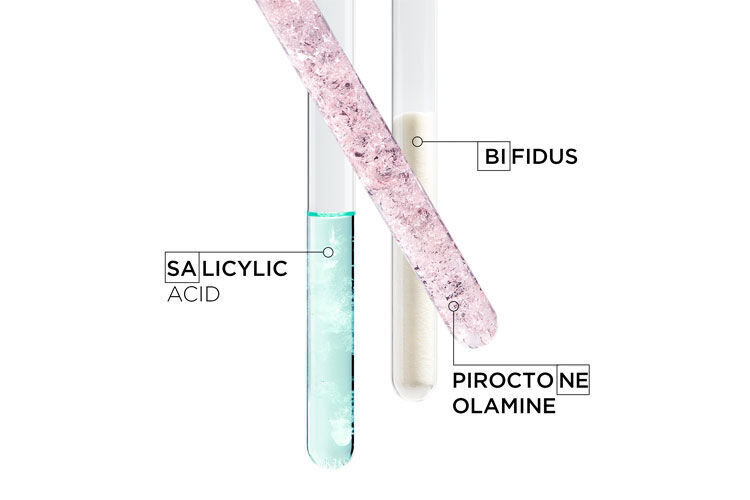 Three test tubes filled with different substances: one contains a pink crystalline solid labeled "Salicylic Acid," the second has a blue liquid labeled "Bifidus," and the third contains a white powder labeled "Piroctone Olamine."