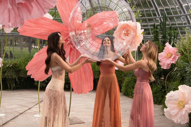 Three women in a greenhouse are joyfully interacting with a large, translucent, bubble-like structure, surrounded by vibrant flower decorations.