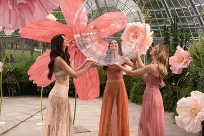 Three women in a greenhouse are joyfully interacting with a large, translucent, bubble-like structure, surrounded by vibrant flower decorations.