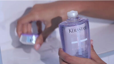 A close-up of a person's hand holding a bottle of Kerastase shampoo, with another product visible in the background.
