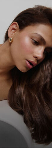 A close-up profile of a woman with long, flowing brown hair, wearing a simple earring, and gazing thoughtfully to the side.