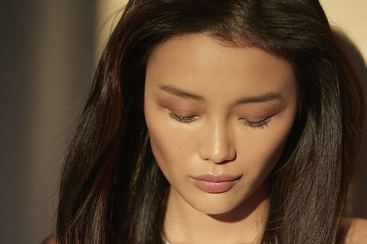 A close-up portrait of a woman with long, dark hair, looking down with her eyes closed and a serene expression. Soft, natural lighting highlights her features.