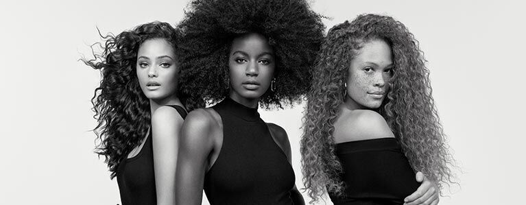 Three women with diverse hairstyles stand side by side against a light background. Each has a unique look: one with voluminous curls, another with an elegant afro, and the third with long, wavy hair. They are wearing stylish outfits and pose confidently.