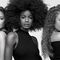 Three women with diverse hairstyles stand side by side against a light background. Each has a unique look: one with voluminous curls, another with an elegant afro, and the third with long, wavy hair. They are wearing stylish outfits and pose confidently.