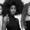 Three women with diverse hairstyles stand side by side against a light background. Each has a unique look: one with voluminous curls, another with an elegant afro, and the third with long, wavy hair. They are wearing stylish outfits and pose confidently.