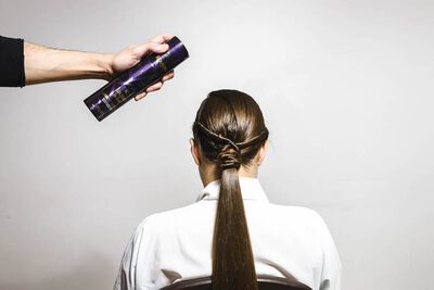 A hand holding a spray can is about to apply hair products to the back of a woman’s long, sleek hairstyle. She is seated and facing away from the camera.