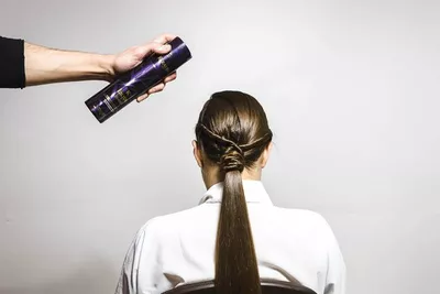 A hand holding a spray can is about to apply hair products to the back of a woman’s long, sleek hairstyle. She is seated and facing away from the camera.