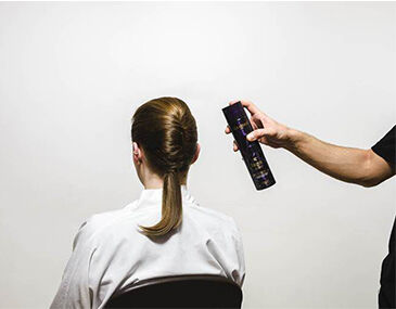 A person with their back to the camera is sitting in a chair while another person is holding a can of hair spray above their head, preparing to style their hair.