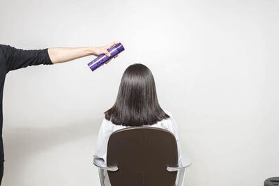A person in a black shirt holds a hair spray can above the head of a seated individual with straight, dark hair, who is facing away from the camera. The background is a plain white wall.