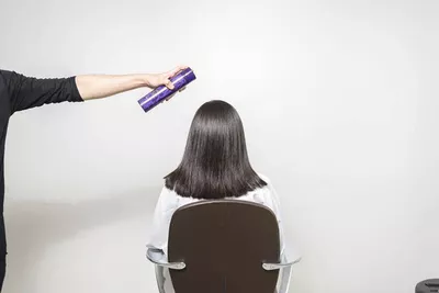 A person in a black shirt holds a hair spray can above the head of a seated individual with straight, dark hair, who is facing away from the camera. The background is a plain white wall.