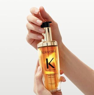 A person’s hands holding a bottle of Kerastase Elixir Ultime hair oil, featuring a gold and black pump design.