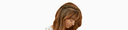 A woman with long, wavy brown hair styled with a side braid, looking downwards against a plain background.