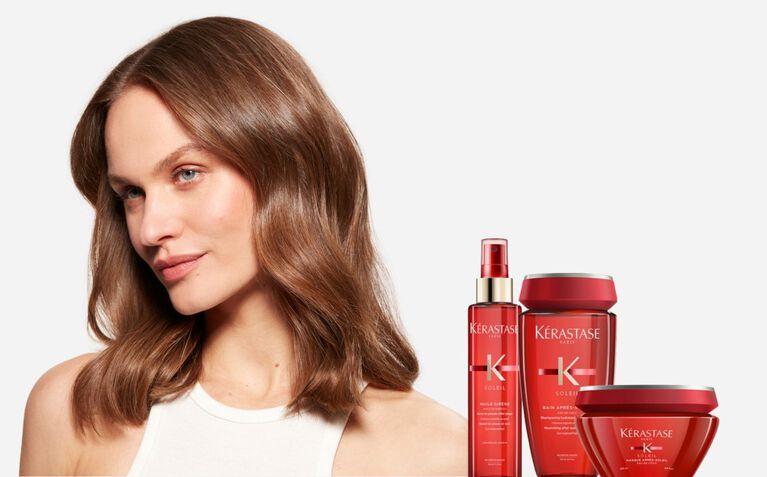 A model with long, wavy hair poses beside a selection of Kerastase hair care products in red packaging.