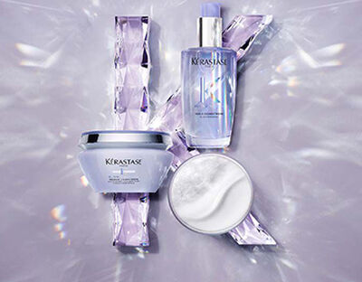 Three Kerastase hair care products arranged on a reflective surface, surrounded by shimmering crystal-like pieces. The items include a jar, a bottle, and a pouch, all featuring soft, pastel colors.