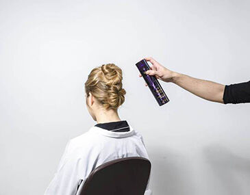 A person with an elegant updo hairstyle sits in a salon chair, while another hand holds a can of hair spray, ready to apply it.