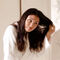 A woman wearing a white robe is brushing her long, dark hair while looking in a mirror. The bathroom has marble accents.