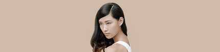 A portrait of a woman with long, dark hair, looking over her shoulder against a neutral beige background. She has a serene expression and is wearing a white sleeveless top.