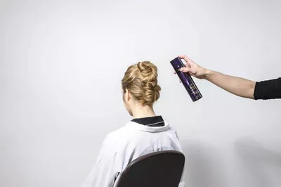 A person with an elegant updo hairstyle sits in a salon chair, while another hand holds a can of hair spray, ready to apply it.