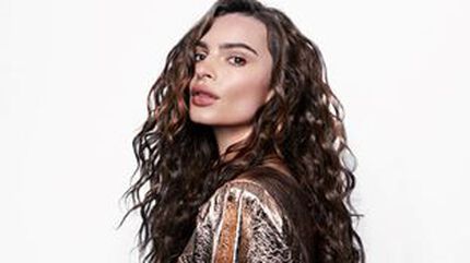 A woman with long, wavy hair looks over her shoulder, wearing a stylish outfit with a textured pattern against a plain white background.