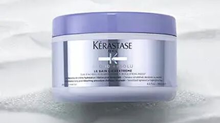 A container of Kerastase hair care product, featuring a sleek design with a lavender lid and a silver label, placed on a textured surface.