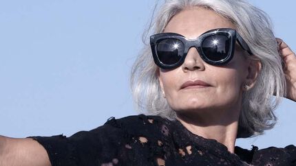 A stylish older woman with silver hair poses confidently outdoors while wearing large, dark sunglasses and a black, distressed top.
