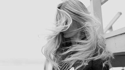 A black and white image of a woman with long, flowing hair, facing away from the camera and partially obscured by her hair. She is positioned near a wooden structure, suggesting an outdoor setting.