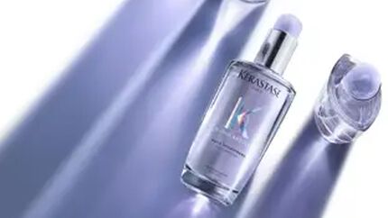 A bottle of Kerastase hair serum surrounded by two smaller bottles, all set against a softly lit background with purple shadows.