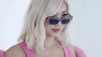 A woman with wavy, shoulder-length blonde hair wears stylish sunglasses and a pink outfit, looking confidently off to the side.