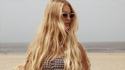 A woman with long, flowing blonde hair wears stylish sunglasses and a checkered top, standing on a beach with calm waves in the background.