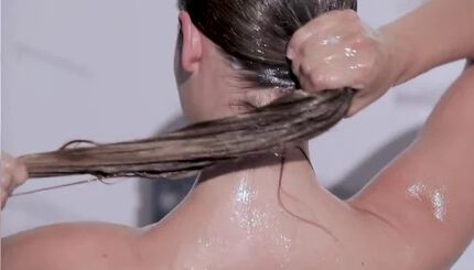 A woman is pulling her long, wet hair into a ponytail from behind, with droplets of water glistening on her shoulders and back.