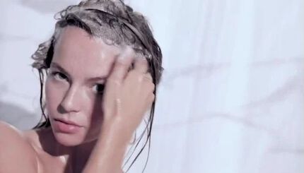 A person with wet hair is applying shampoo in a shower, with water cascading down their face and shoulders. The background features a light-colored, textured surface.