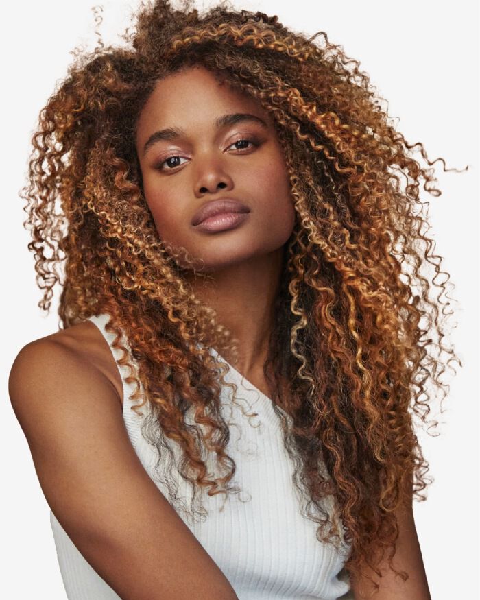 A close-up portrait of a young woman with voluminous, curly hair and a natural makeup look, wearing a sleeveless top. She has a confident expression.